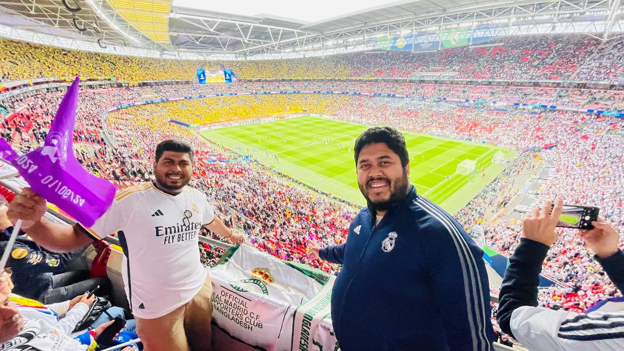 Two PMBD members holding the club's official banner and a purple flag in the stands at Wembley Stadium, packed crowd and pitch visible before a Champions League Final